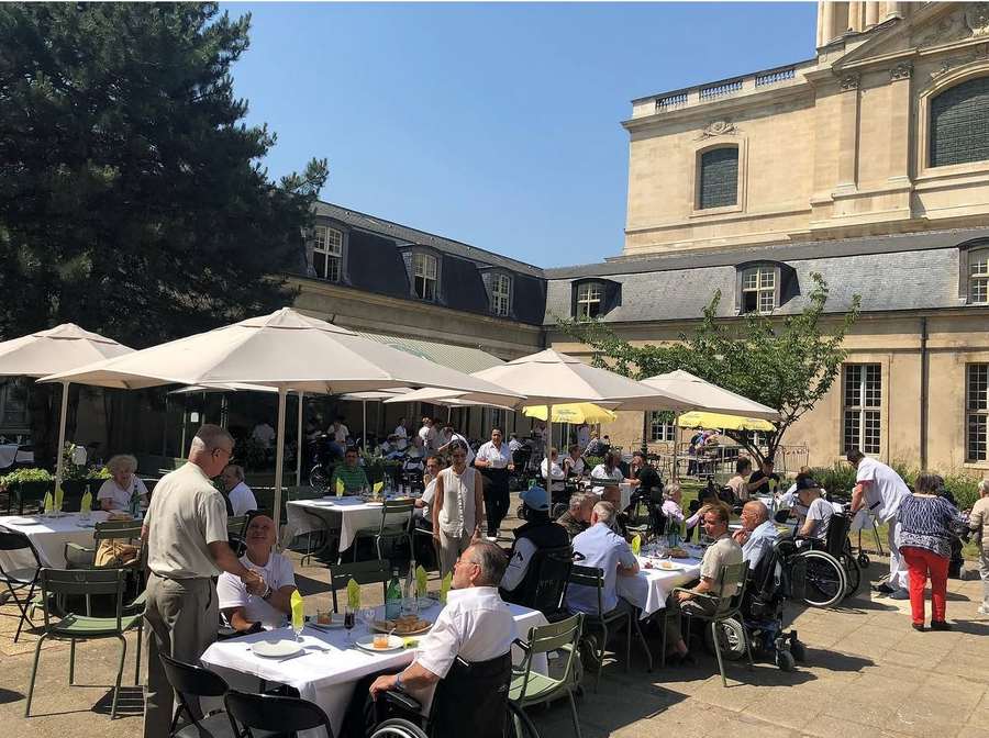 Déjeuner en terrasse dans la cour de l'INI : pensionnaires, familles et personnel réunis sous les parasols, façade des Invalides en arrière-plan