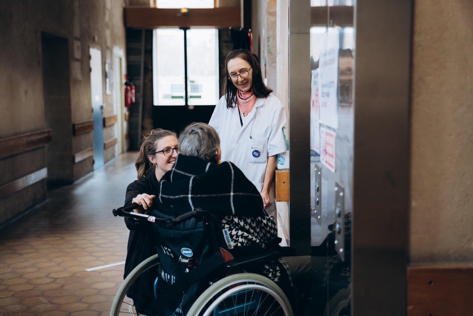 Deux soignantes de l'INI échangent chaleureusement avec un pensionnaire en fauteuil roulant dans un couloir de l'Institution