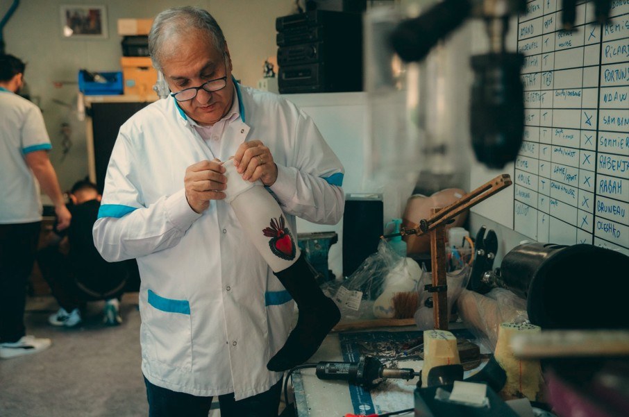 Un prothésiste de l'INI, en blouse,réalise à la main la finition d'une prothèse noire ornée d'un habillage brodé, dans l'atelier d'appareillage
