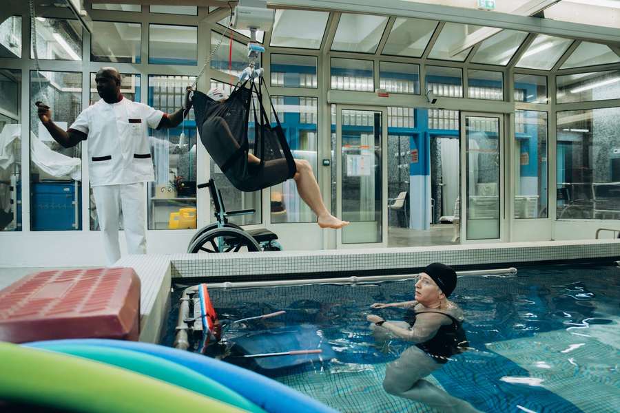 Un soignant de l'INI descend un patient dans le bassin de balnéothérapie à l'aide d'un lève-personne, tandis qu'une patiente nage à proximité