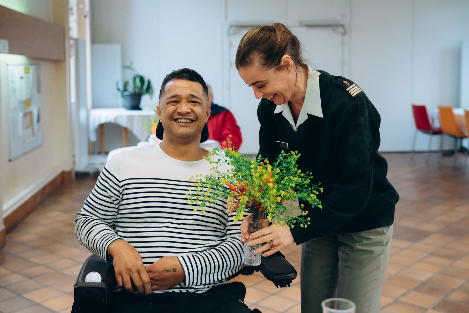 Une soignante en uniforme offre un bouquet à un pensionnaire de l'INI en fauteuil roulant, tous deux souriants dans un espace de vie de l'Institution