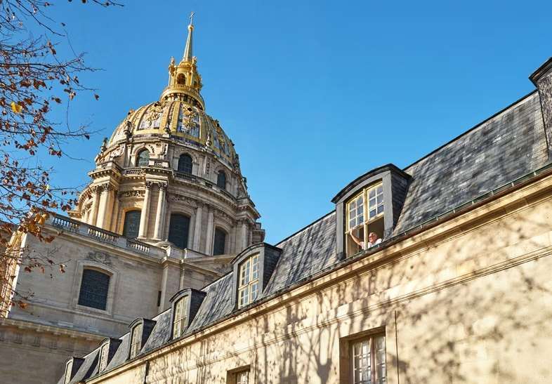 Le dôme doré des Invalides vu au-dessus des toits sous un ciel d'automne, une personne saluant depuis une lucarne